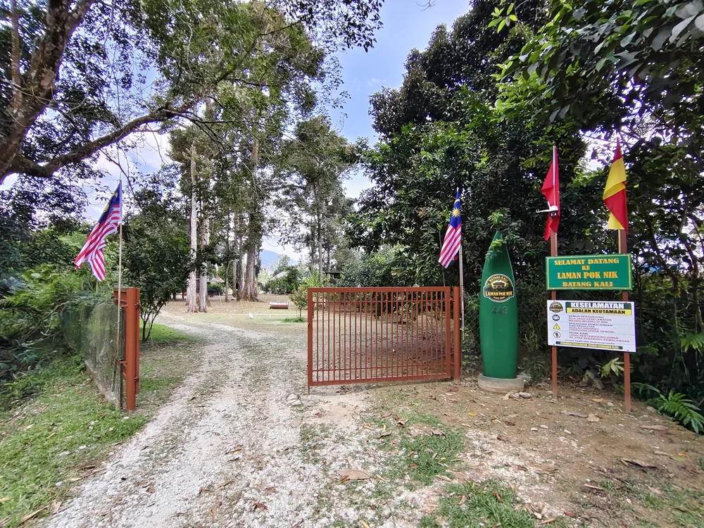 Pintu masuk Laman Pok Nik: pagar besi perang, penanda berbentuk surfboard warna hijau bernombor 443 dan berlogo Laman Pok Nik, serta papan tanda Selamat Datang dan Keselamatan, dengan jalan berbatu di hadapan