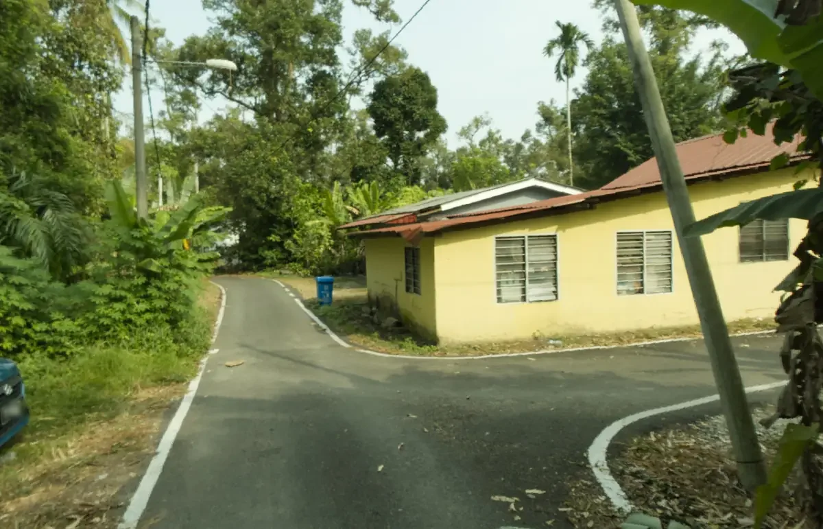 Jalan bertar membelok melepasi rumah setingkat berwarna kuning berbumbung merah, pada aras mata pemandu